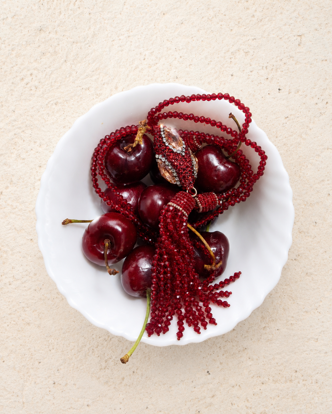 A white ceramic bowl filled with glossy dark red cherries and draped with deep red beaded strands and a tassel ornament, set on a textured beige surface.