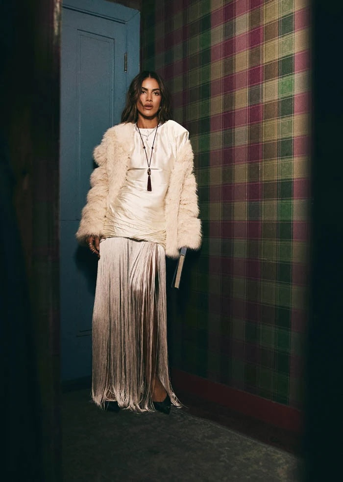 A model stands in a dimly lit hallway wearing a cream-colored faux fur jacket over a matching cream top and a long pleated metallic skirt. The background features a blue door on the left and a green and purple plaid wallpaper on the right.