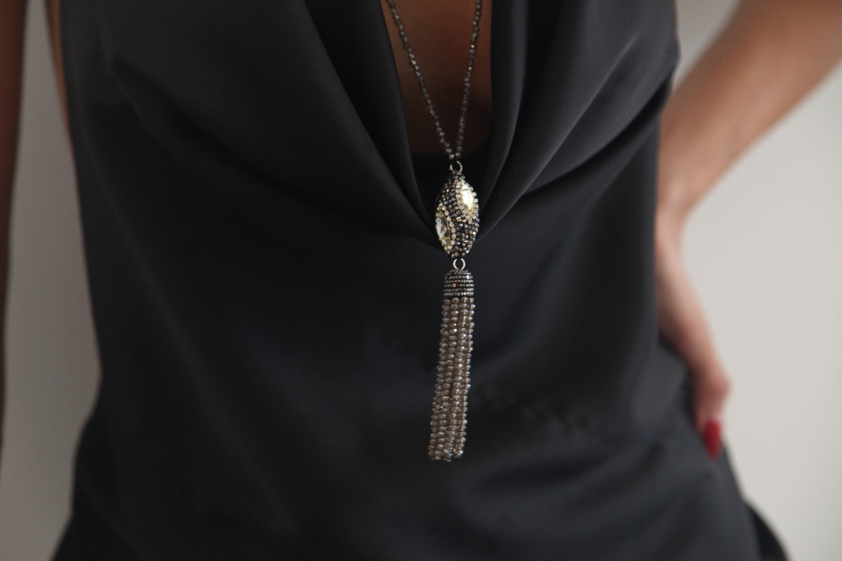A close-up of a model wearing a long silver-toned tassel necklace with a textured oval pendant over a black cowl-neck top.