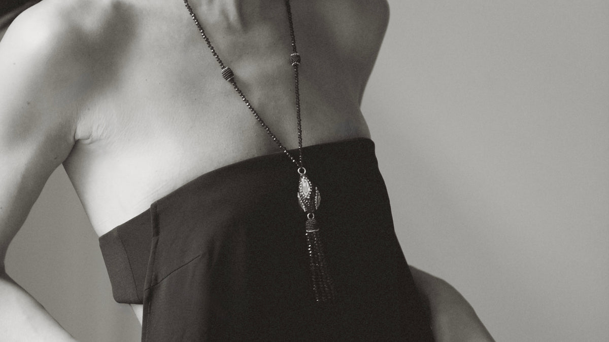 A close-up black and white photo of a model wearing a long dark chain necklace with a decorative pendant and tassel, paired with a strapless dark dress.