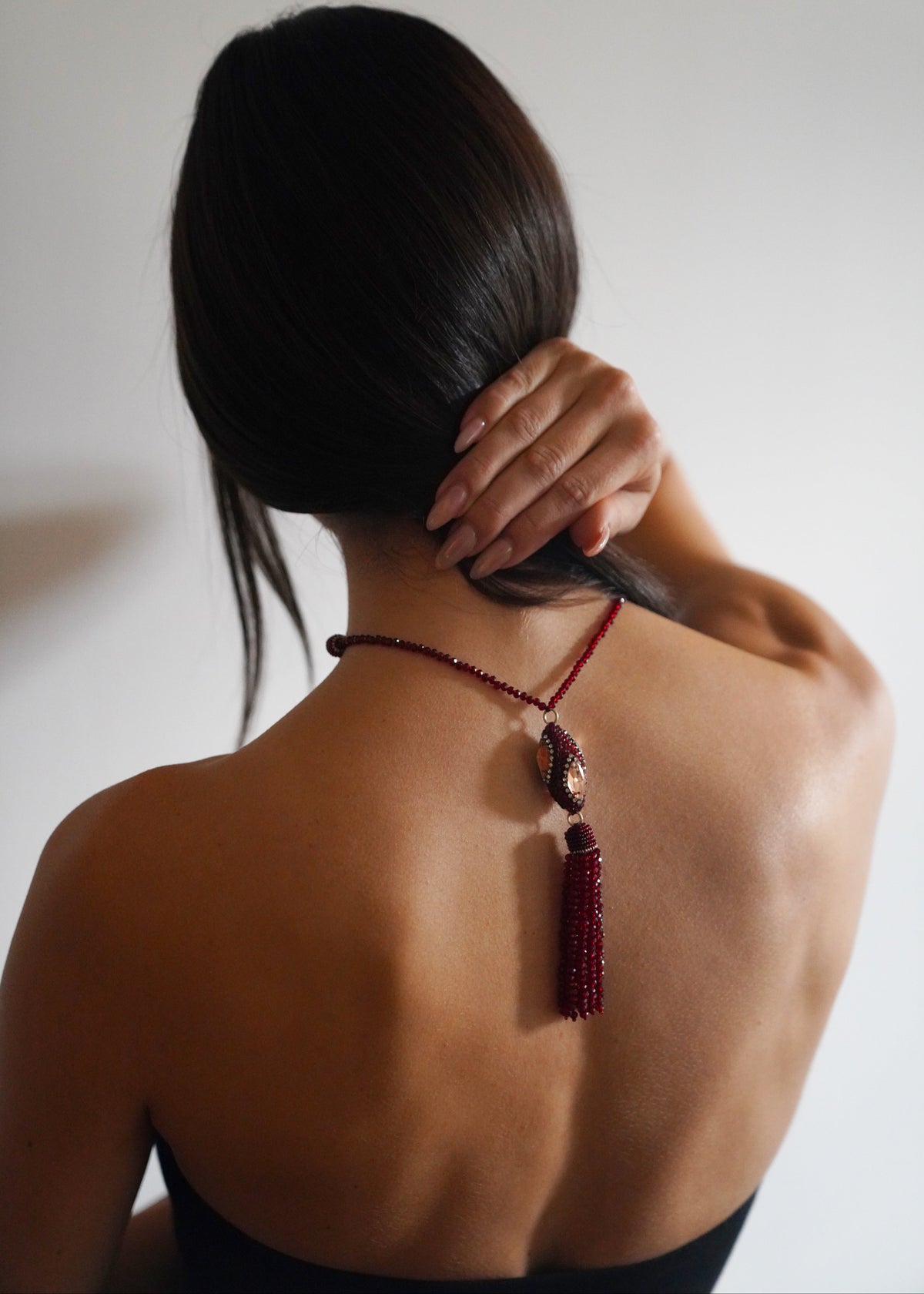 A woman shown from the back wearing a dark red beaded necklace with a tassel and central pendant, set against a plain light gray background.