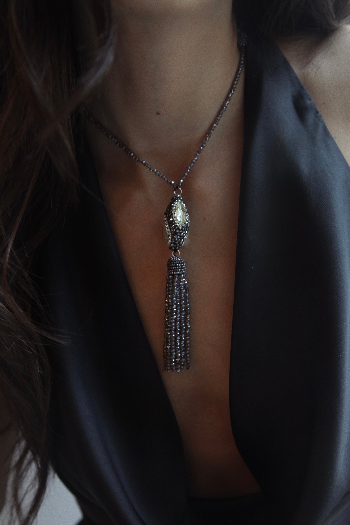 A close-up of a model wearing a dark silver tassel necklace with a teardrop-shaped crystal pendant, styled with a black satin top.