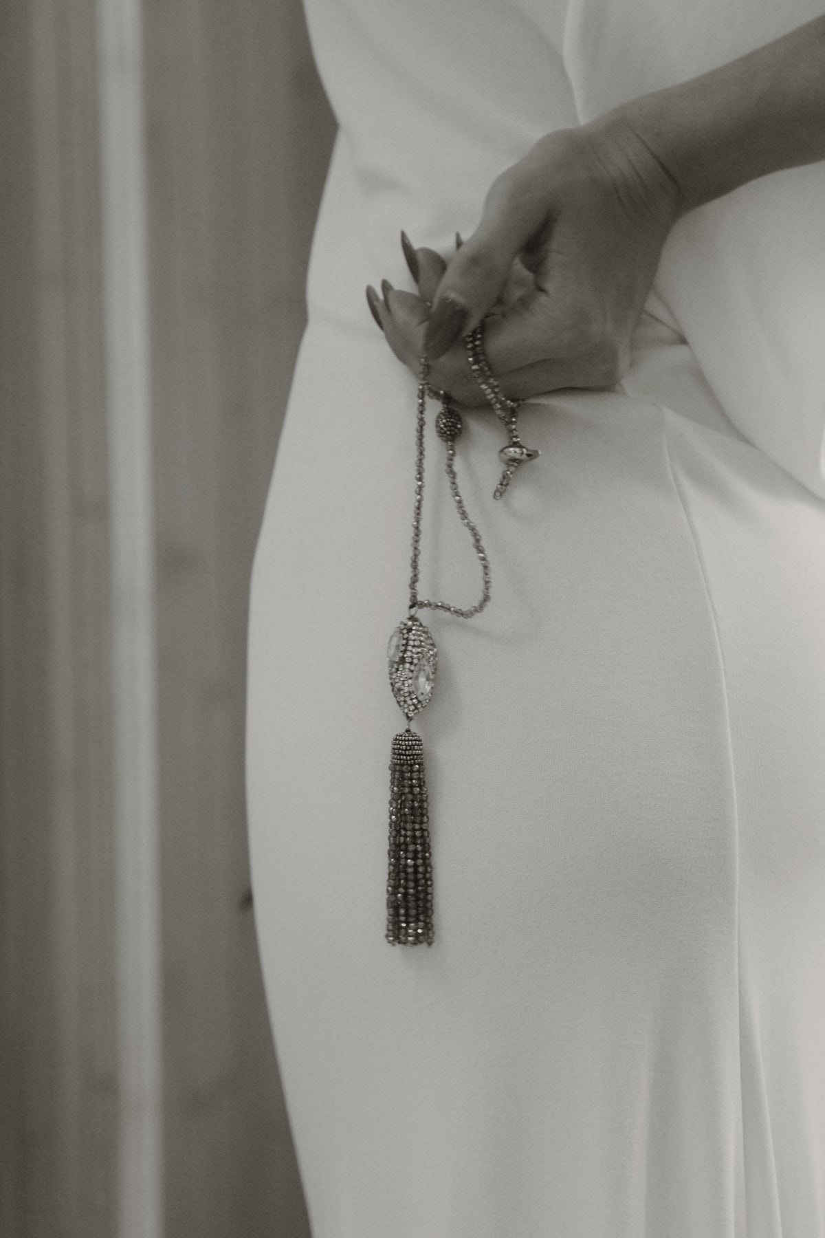 A close-up of a person in a white dress holding a long beaded tassel necklace with a teardrop-shaped crystal pendant.