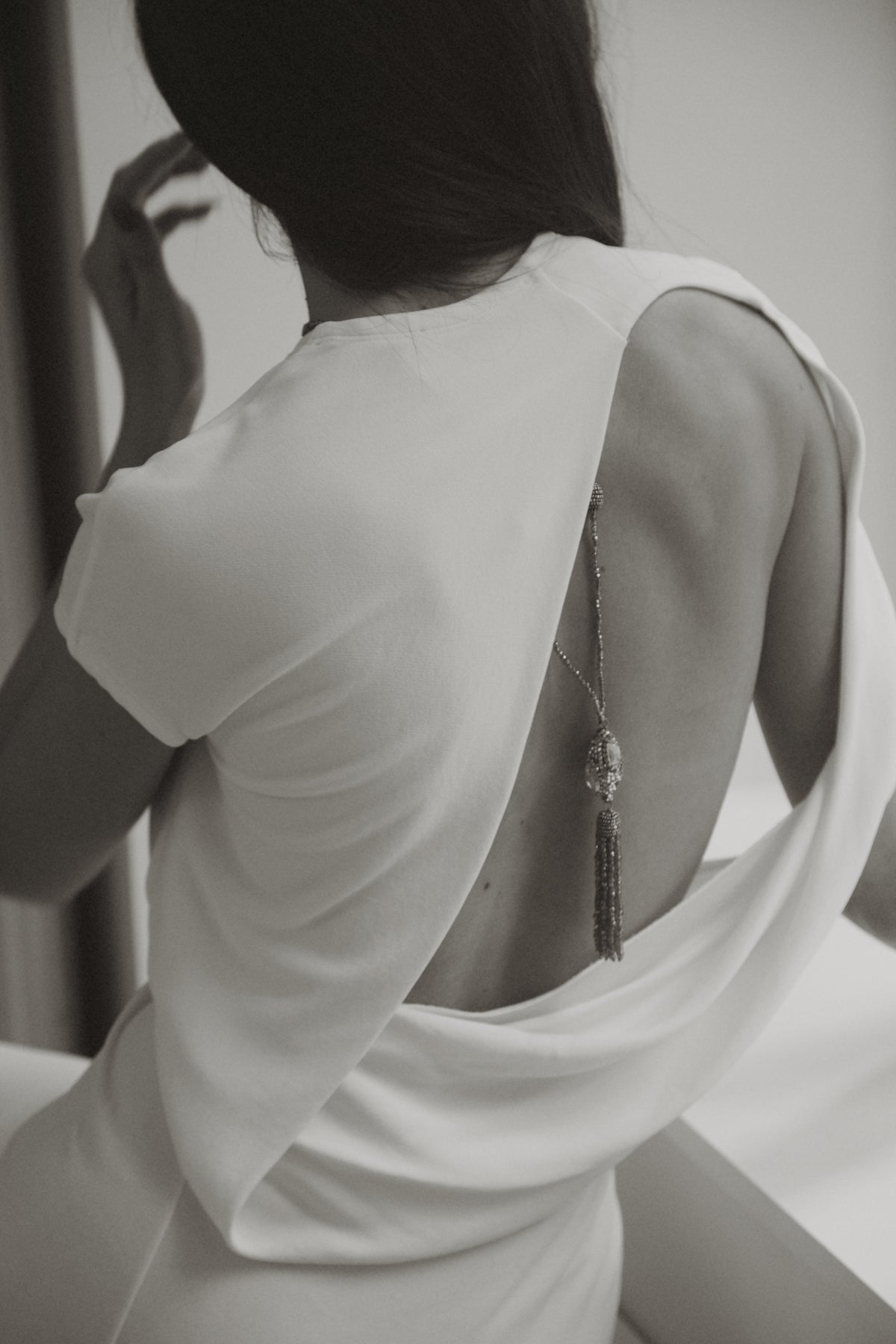 A woman shown from behind wearing a white sleeveless dress with a large open back and a long dangling tassel necklace resting on her back. The photo is in black and white.